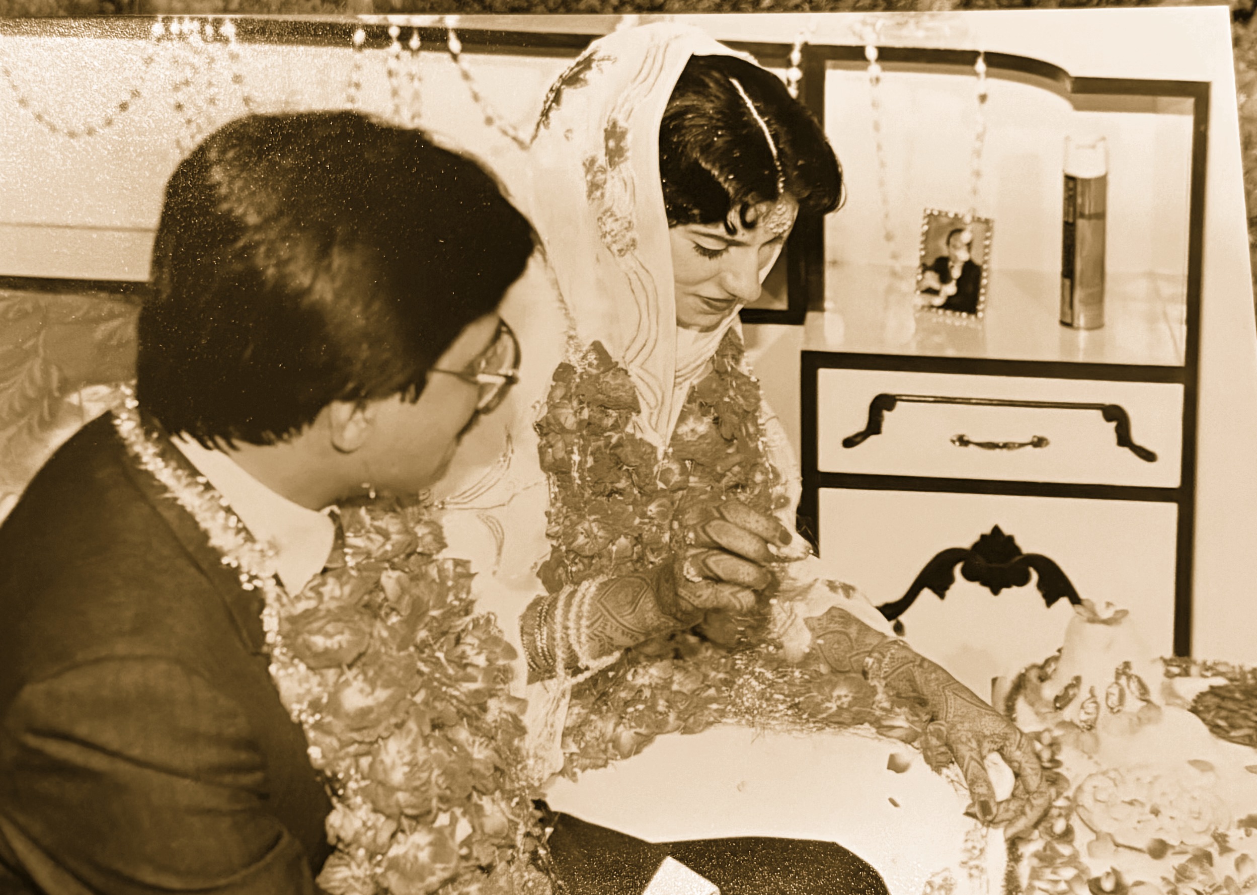A vintage family photo from a wedding in Pakistan, a bride with henna-decorated hands and a groom in a floral garland sharing a sweet dessert